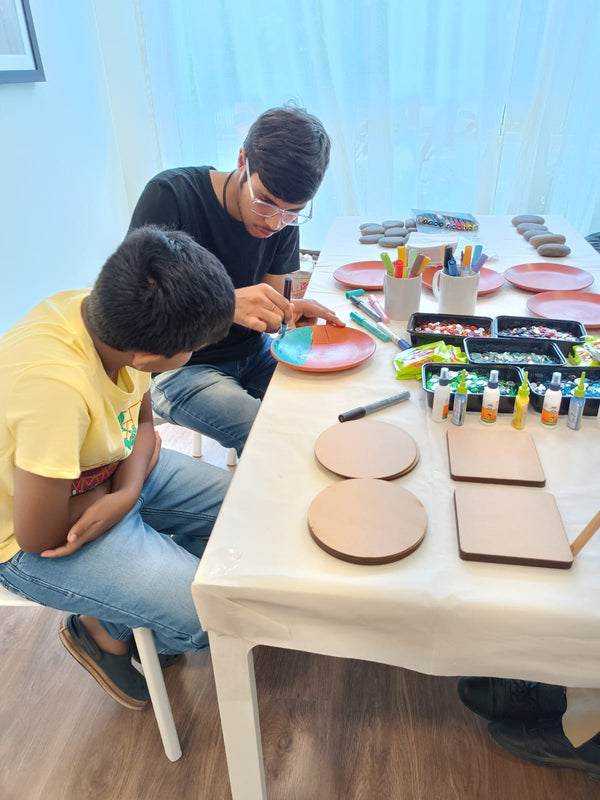 Step 3: two guests painting coasters at a Kitsters station, finished pieces beside them