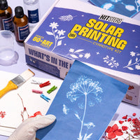 Solar printing kit in the backgroud, showing a hand holding a cyanotype print of a dandelion, with cyanotype chemicals, wooden brush, wooden clips, a beaker and pressed flowers in the background