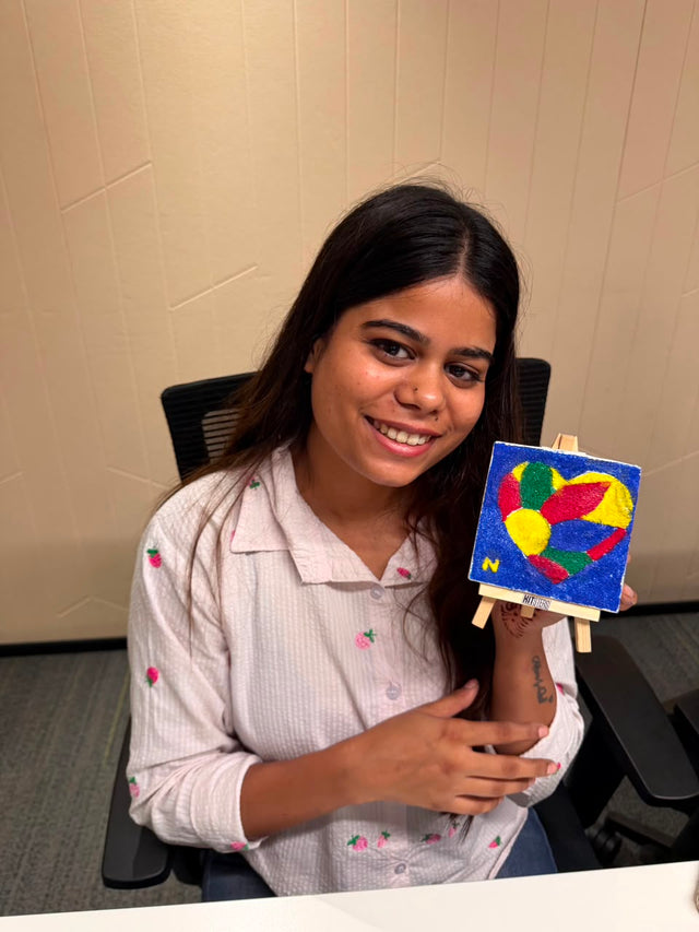 A colleague at her desk holding up a mini canvas painted in bright red, yellow, blue and green on its easel