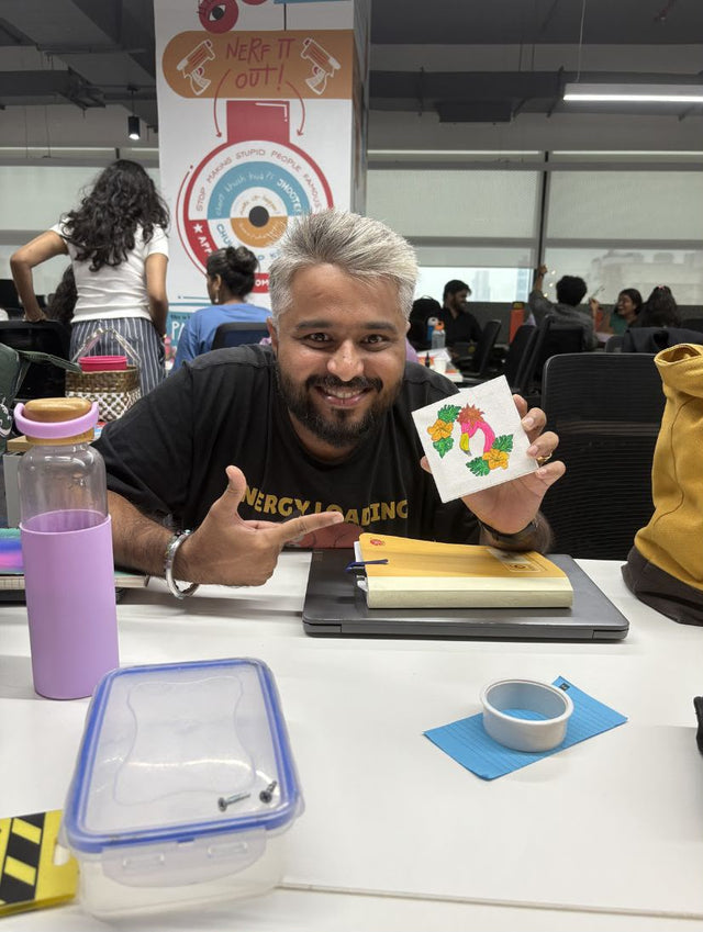 A colleague at a long office table holding up a small painted canvas, kit materials in front of him