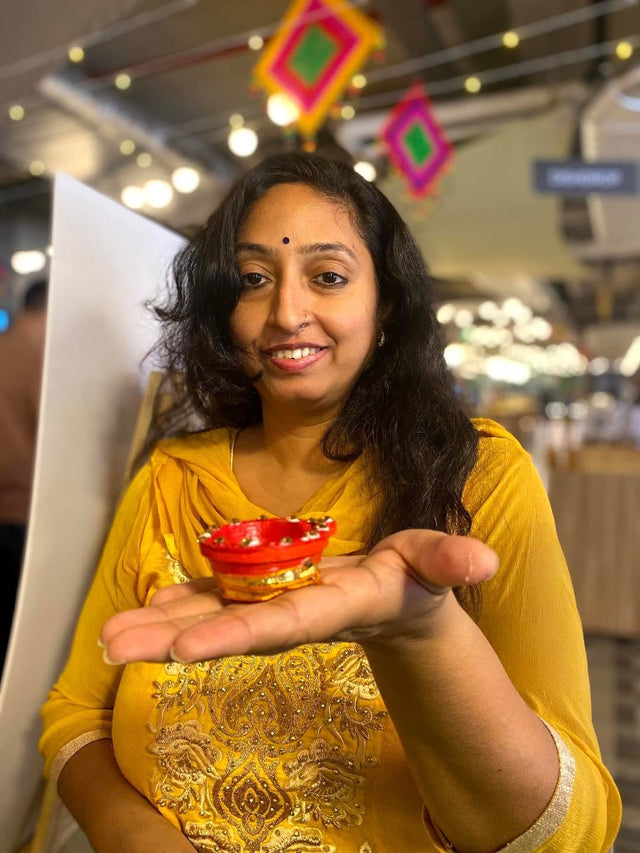 A colleague in a yellow kurta holding a freshly painted red and gold diya on her open palm